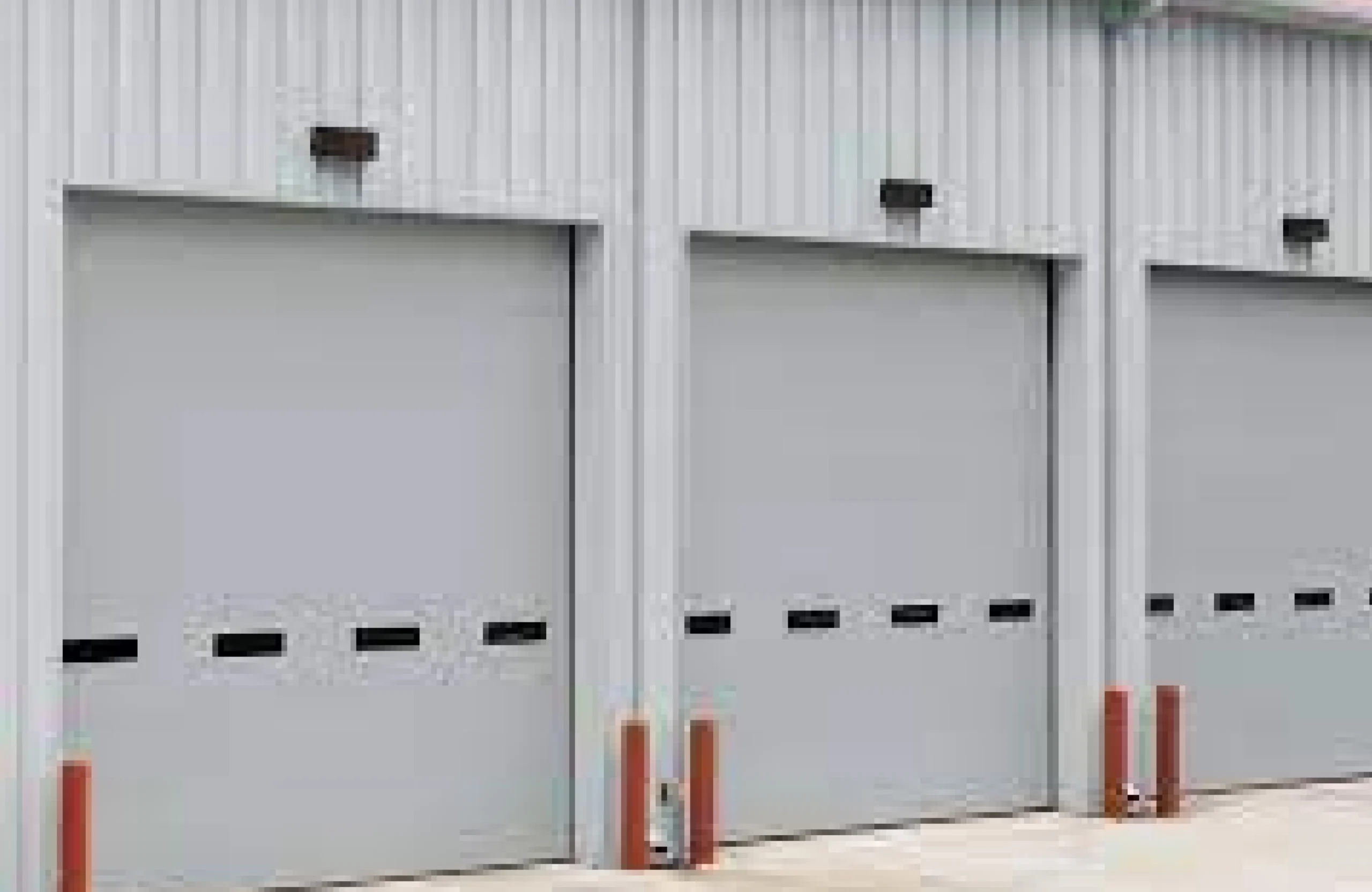 Three commercial sectional doors with vision panels and protective bollards on a metal clad industrial unit.