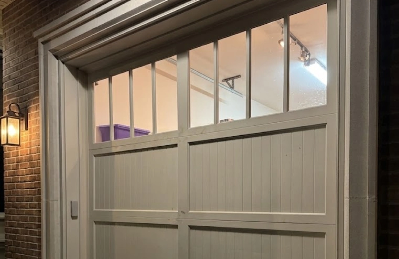 Cream carriage house style garage door with vertical plank panels and divided glass windows on a brick home.