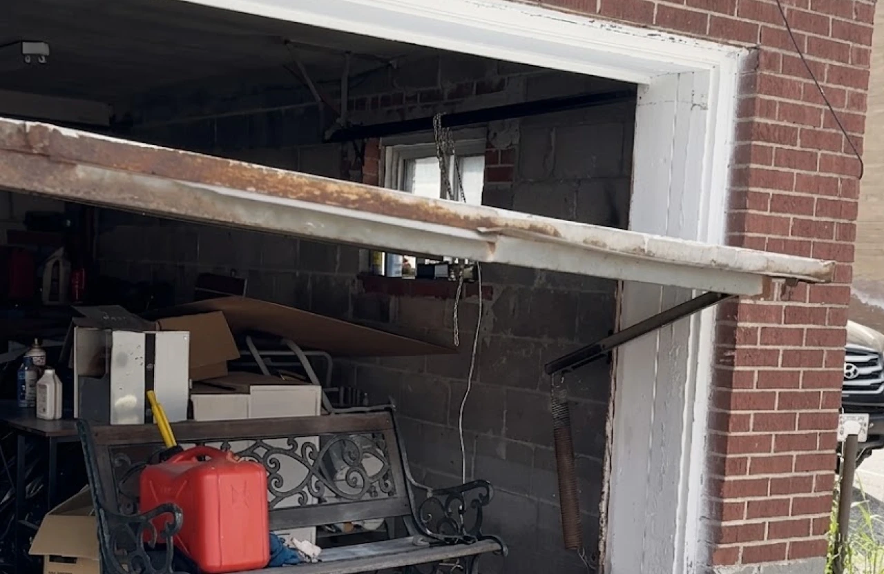 Older tilt-up garage door stuck at an angle in a brick garage opening, dropped lower on one side.