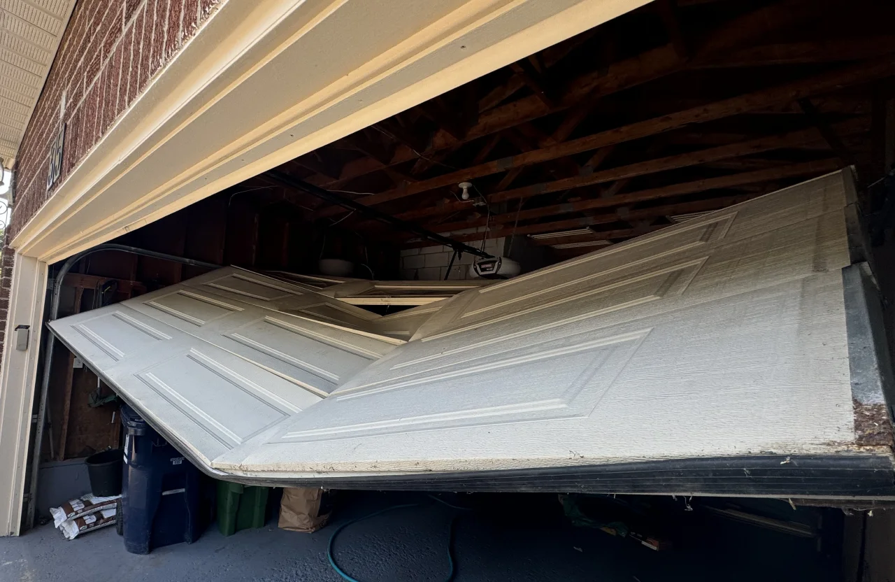 Garage door off track with panels buckled and collapsed inward, hanging at an angle across the garage opening.
