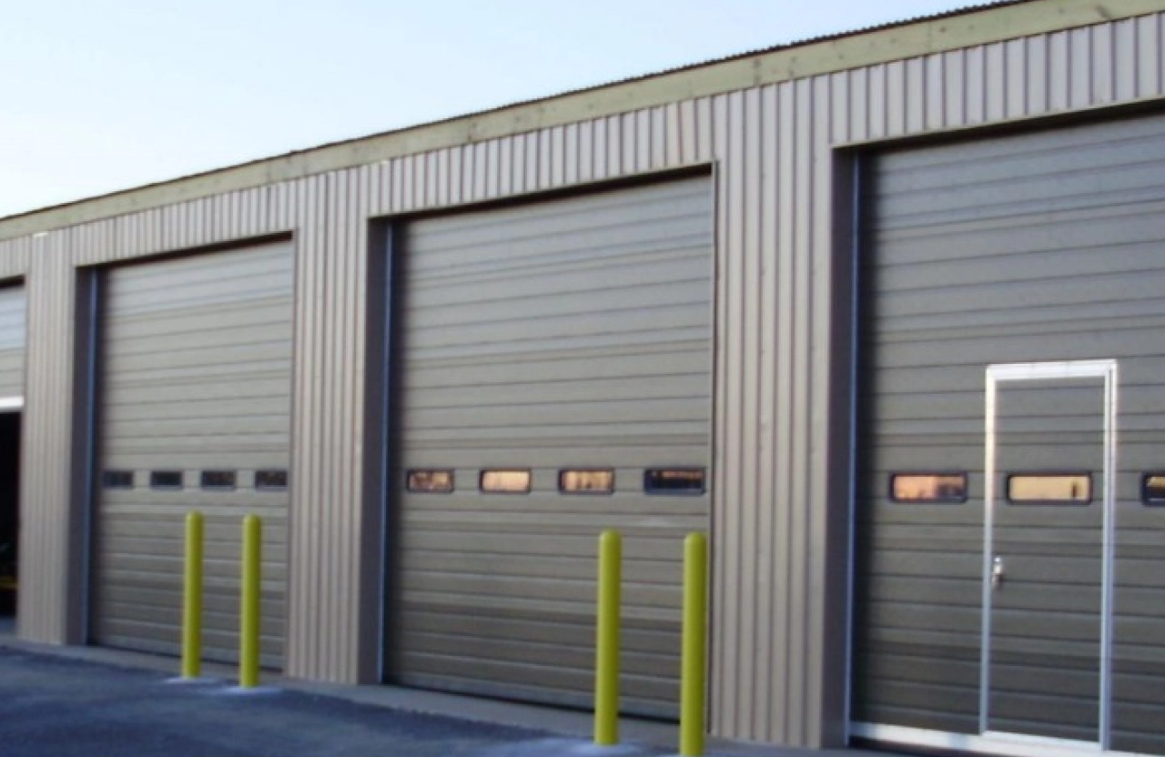 Row of industrial overhead doors with vision panels and yellow bollards on a metal clad warehouse building.